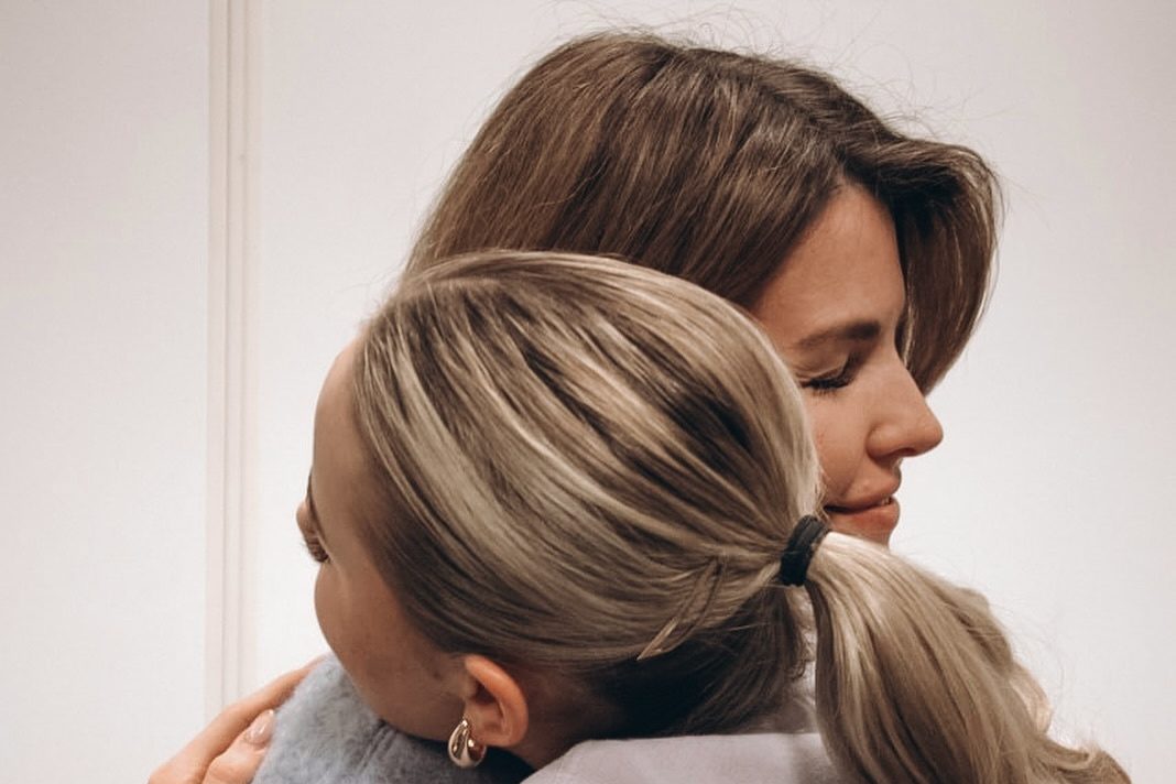 Zwei Frauen umarmen einander vor einer hellen Wand.
