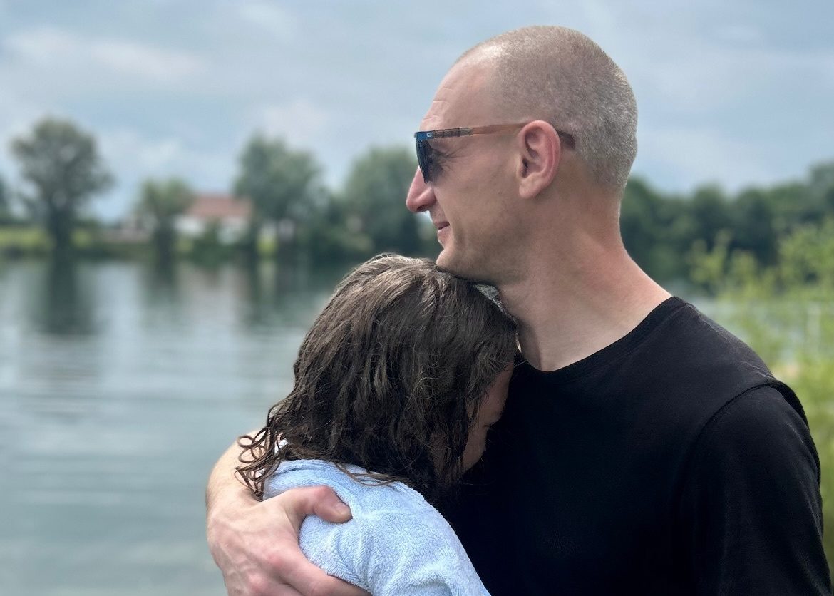 Ein Mann hält am Seeufer eine Person im Arm, die in ein hellblaues Handtuch gehüllt ist.