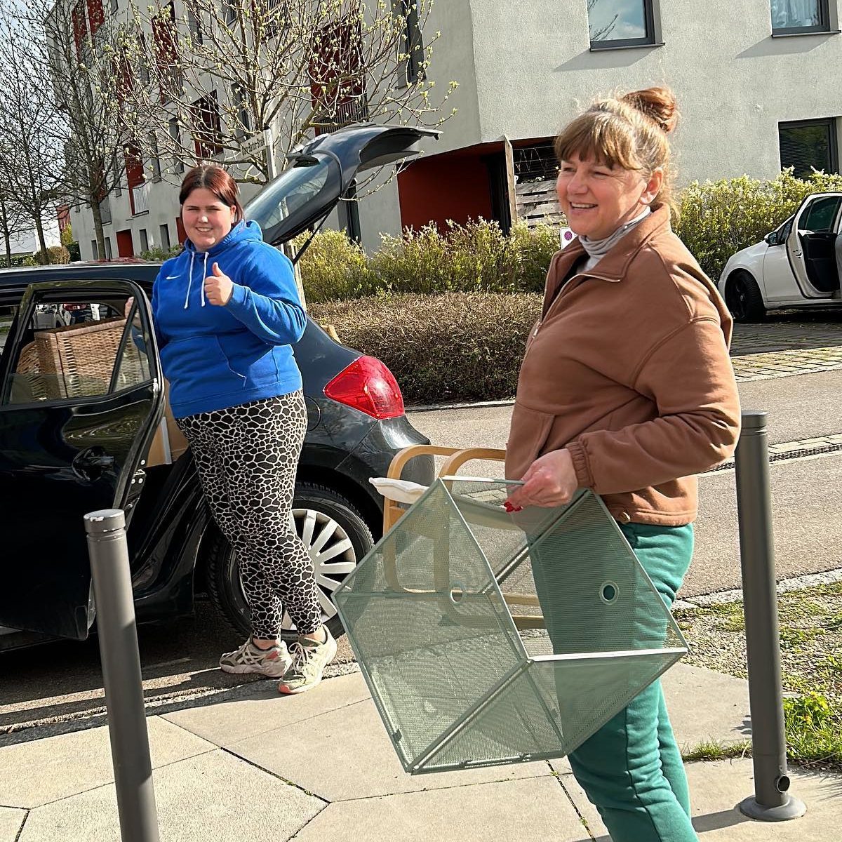 Zwei Frauen laden vor einem Wohnhaus ein Auto aus, eine trägt einen Gitterkorb.