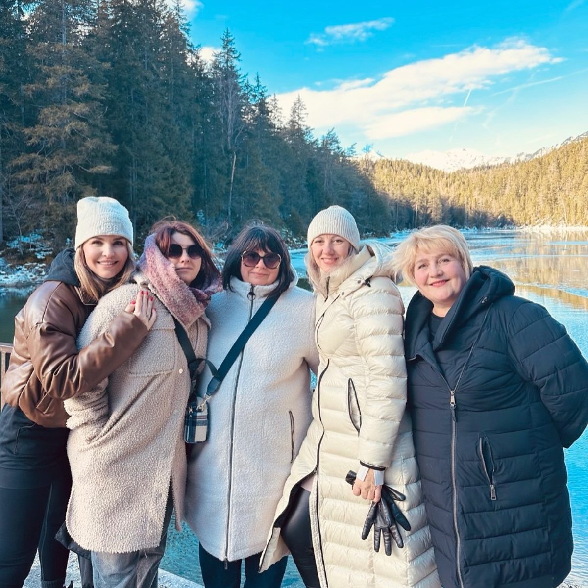 Fünf Frauen in Winterjacken stehen an einem Bergsee unter blauem Himmel.