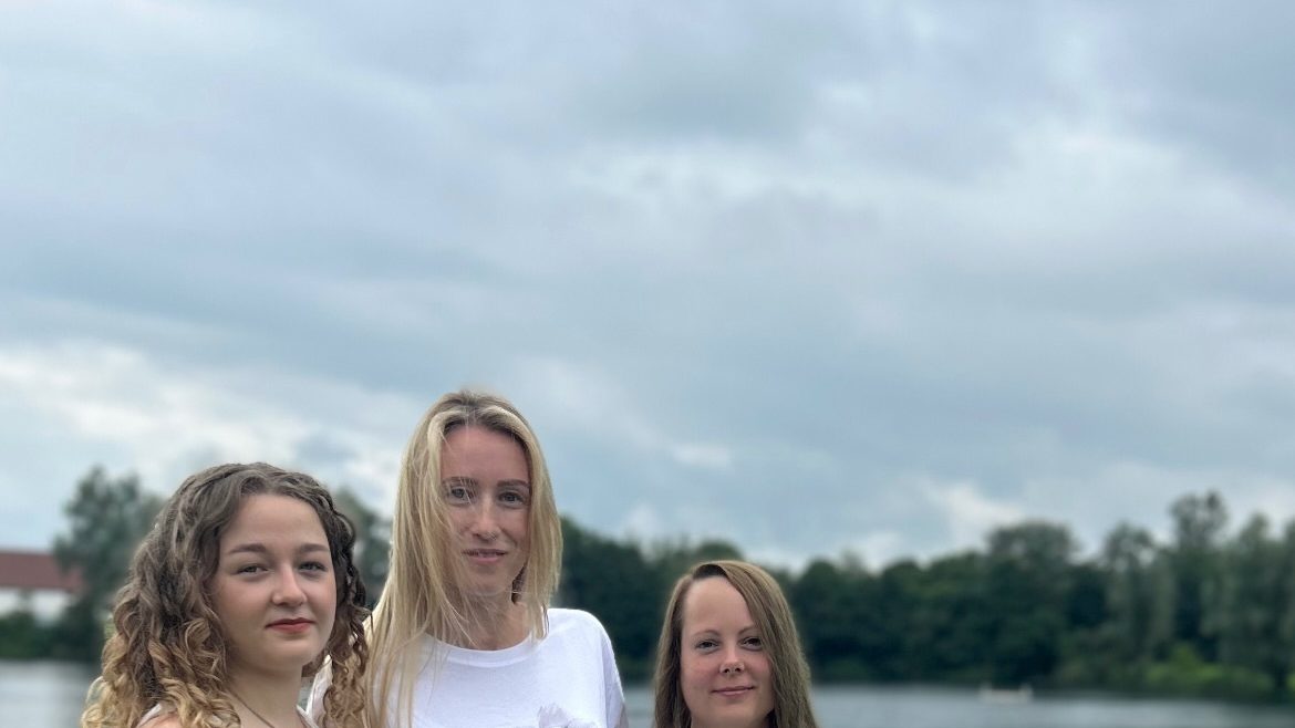 Drei Frauen in weißer Kleidung stehen auf einer Wiese am See, über ihnen ein weiter Wolkenhimmel.