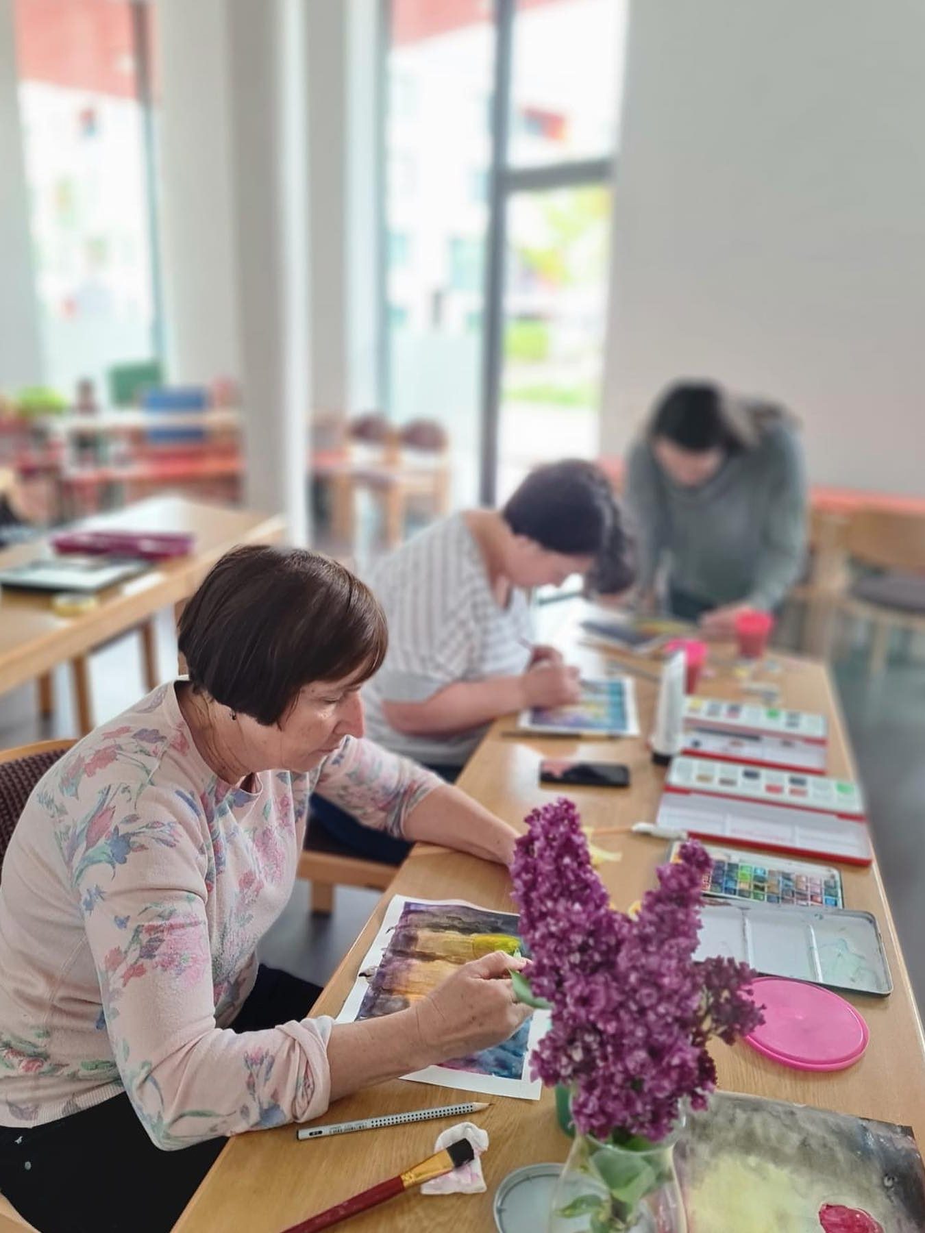 Drei Frauen malen an einem langen Holztisch mit Aquarellfarben, vorn steht ein Fliederstrauß.