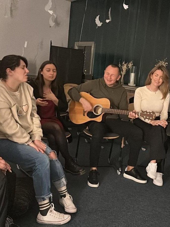Vier Menschen sitzen im Kreis und singen mit geschlossenen Augen, einer spielt Gitarre.