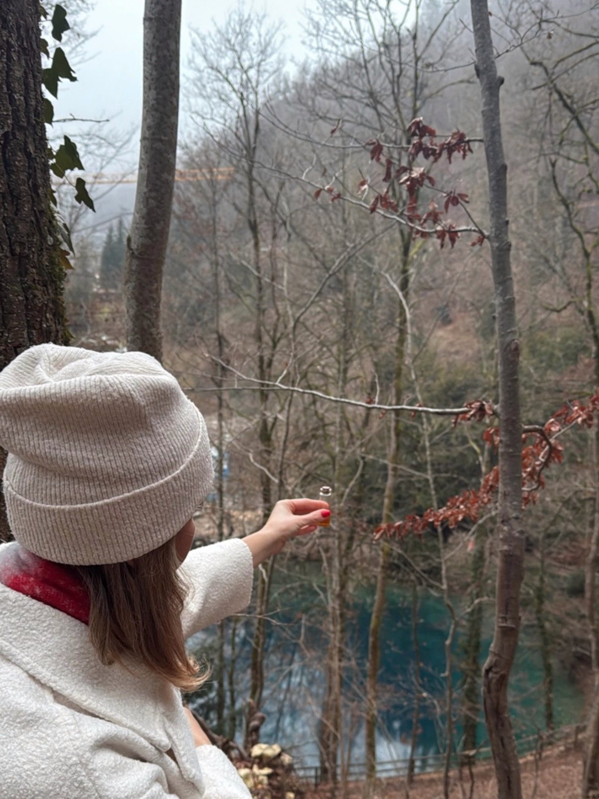 Eine Frau mit heller Mütze, von hinten gesehen, streckt die Hand über türkisblauem Wasser im Winterwald aus.